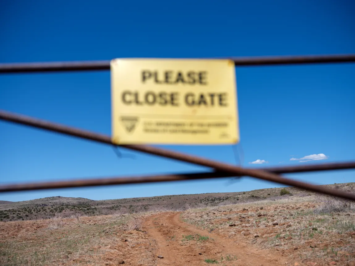 Un panneau jaune flou indiquant « Veuillez fermer la porte » est monté sur un grillage au premier plan. À travers la clôture, une vue nette révèle un chemin de terre sinueux traversant un paysage sec et vallonné sous un ciel bleu vif.