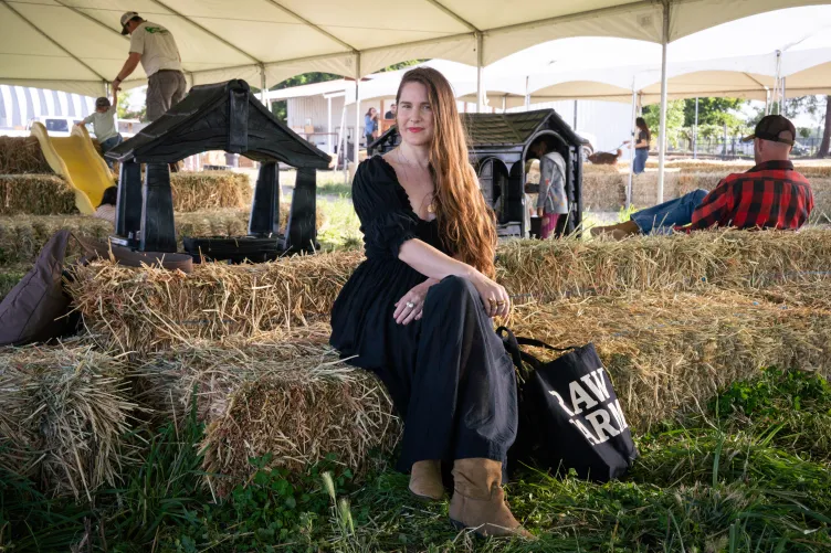 Une femme vêtue d’une robe noire est assise sur une botte de foin à côté d’un sac fourre-tout Raw Farm sous une tente blanche, avec d’autres personnes en arrière-plan.