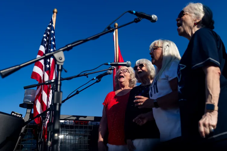 Quatre femmes se tiennent côte à côte et chantent dans des microphones en plein air lors d'un événement en journée. Des drapeaux les flanquent en arrière-plan sur un ciel bleu clair et profond.