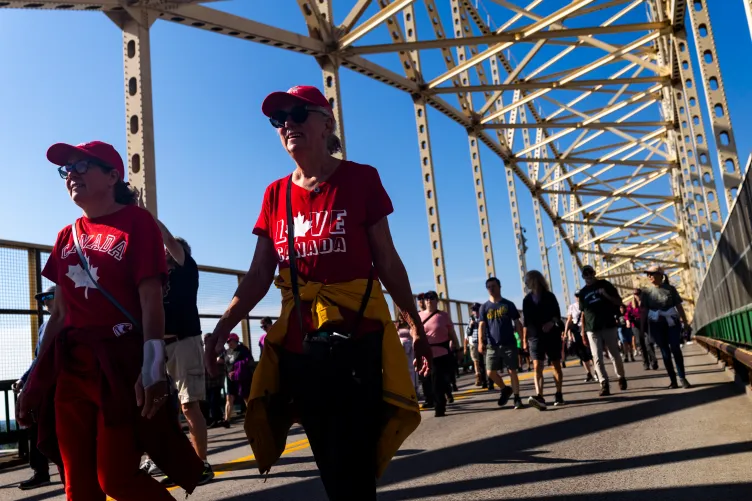 Deux femmes portant des chemises rouges assorties au thème du Canada et des casquettes rouges traversent un pont au milieu d'une foule de piétons. Le cadre du pont ensoleillé se dresse au-dessus d'un ciel bleu clair.