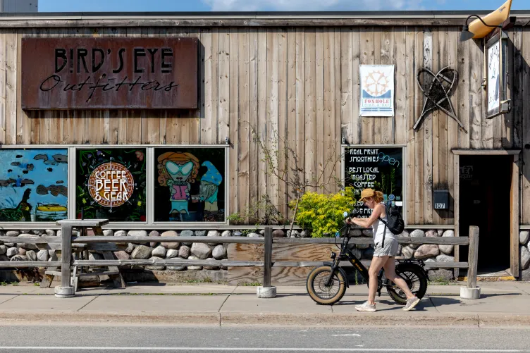 Une femme portant une casquette beige, un débardeur blanc, un short et un sac à dos pousse un vélo électrique noir le long d'un trottoir devant une devanture rustique en bois. Le bâtiment comporte une enseigne métallique indiquant « Bird's Eye Outfitters » au-dessus des fenêtres décorées d'œuvres d'art et de textes annonçant le café, la bière, les équipements et les smoothies.