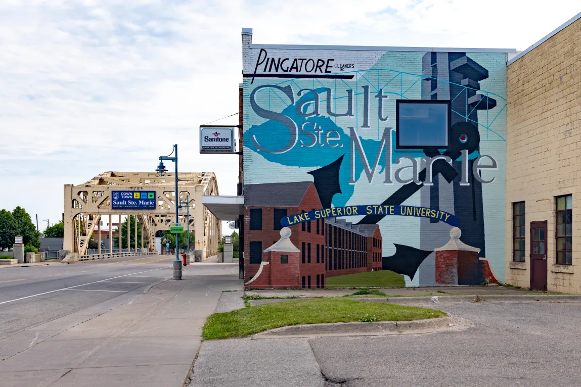 Une vue au niveau de la rue montre un bâtiment orné d'un grand Sault Ste. Marie murale qui comprend les expressions « Pingatore Cleaners Inc. » et «Université d'État du Lac Supérieur». En arrière-plan, un pont en acier jaune clair enjambe une route sous un ciel partiellement nuageux.