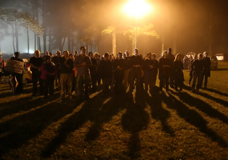Une foule de gens se tient devant des bois ombragés et un lampadaire jaune, projetant des ombres allongées sur la pelouse devant eux.