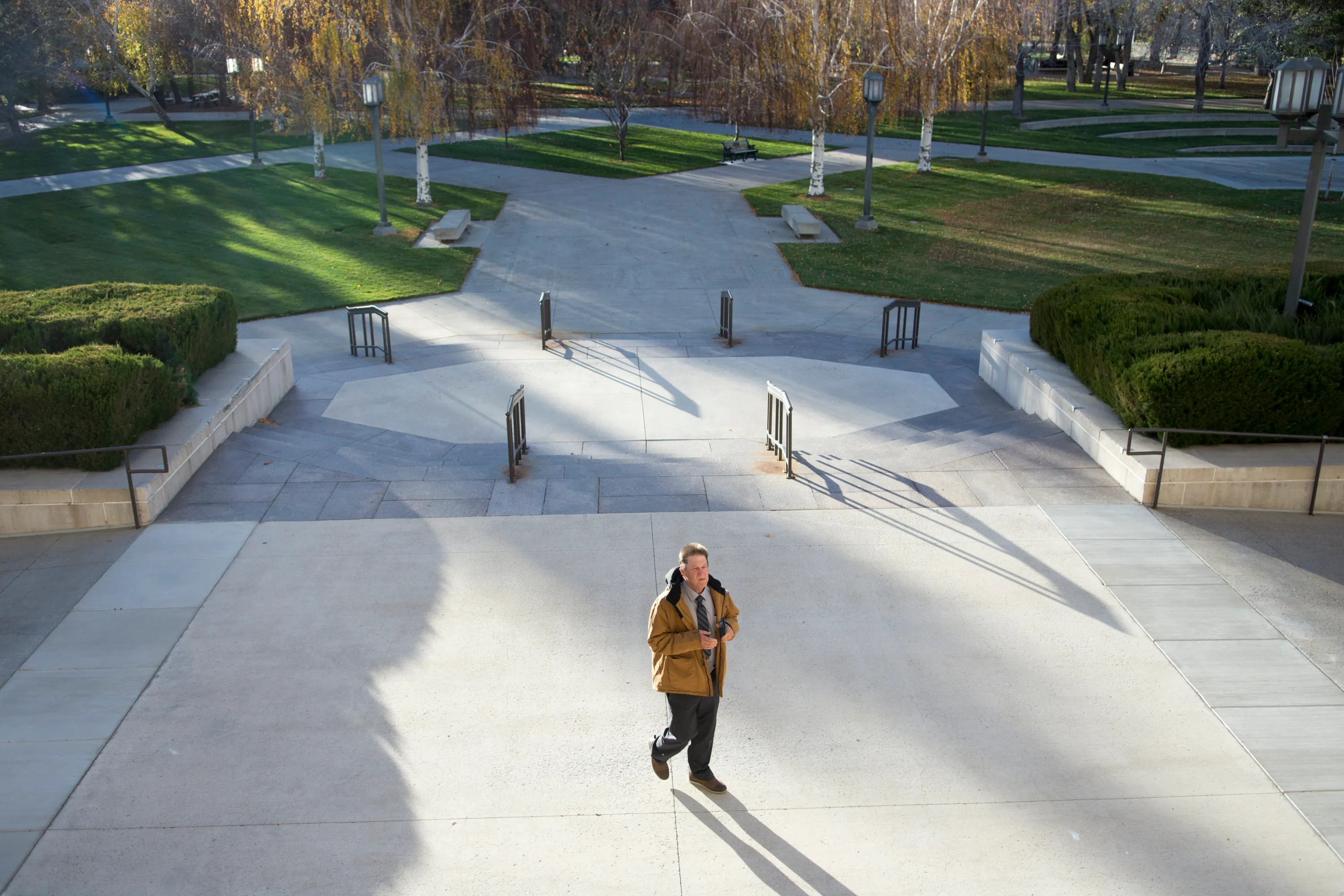 Une vue aérienne d'un grand escalier avec des pelouses et des arbres bien entretenus. Un homme portant un manteau moutarde et une cravate grise se tient au centre, avec son ombre projetée devant lui.