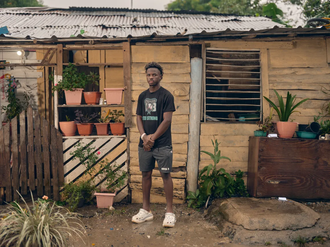 Un jeune homme portant un T-shirt noir et des sabots blancs se tient devant une maison rustique en bois entourée de plantes en pot.
