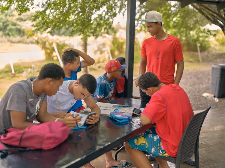 Un groupe de jeunes joueurs de baseball masculins sont assis autour d’une table de pique-nique sombre sous un abri extérieur couvert, étudiant, écrivant dans des cahiers et regardant des téléphones.