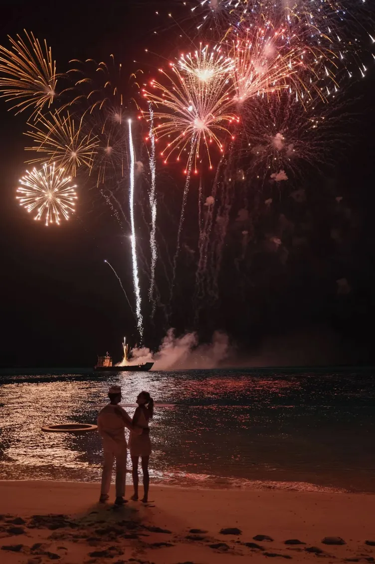 Un homme et une femme se tiennent sur une plage alors que des feux d’artifice explosent au-dessus de l’eau.