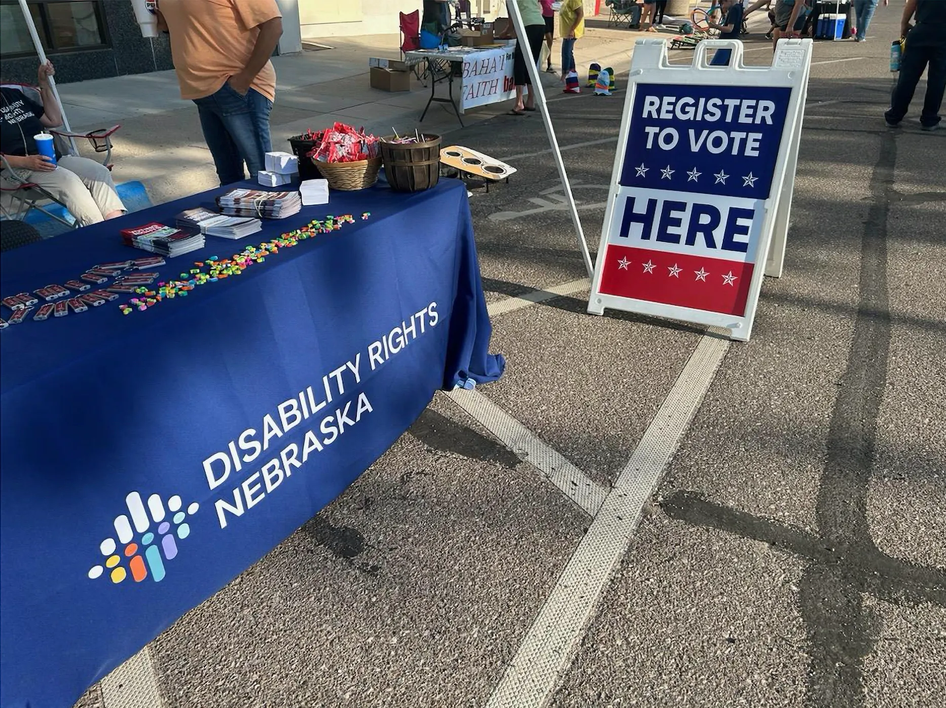 Une table avec une bannière bleue indiquant « Disability Rights Nebraska » se trouve sur le côté gauche de l'image tandis qu'un panneau sandwich rouge, blanc et bleu demande aux gens de s'inscrire pour voter à la table.