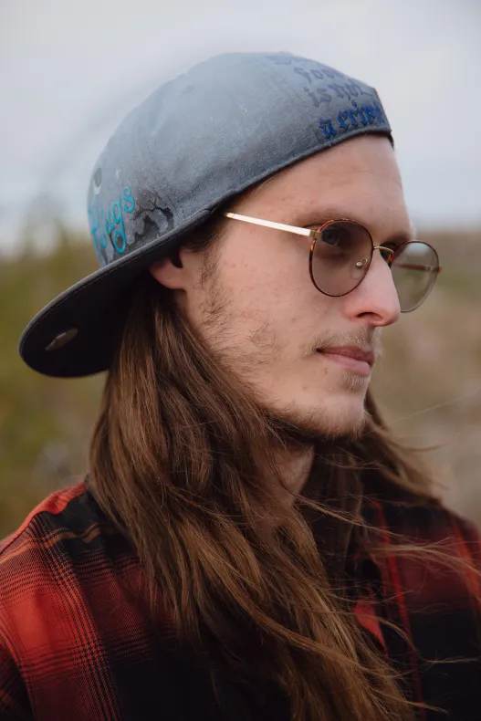 Portrait en gros plan d'un homme aux cheveux longs, aux lunettes rondes et à la casquette de baseball grise avec un texte manuscrit.