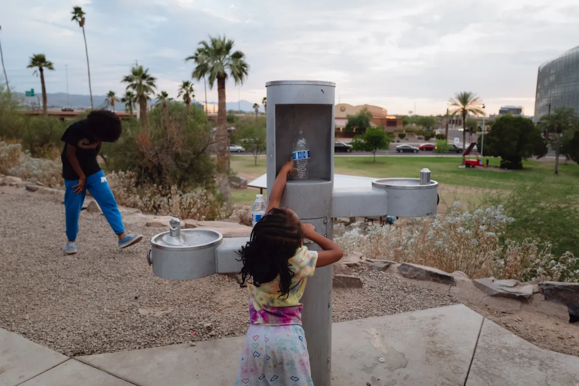 Une jeune fille tend la main vers une bouteille d’eau placée dans une station d’hydratation d’un parc public tandis qu’un autre enfant joue à proximité.