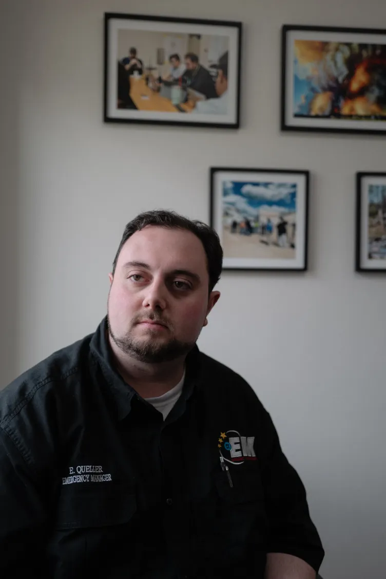 Un homme est assis à l’intérieur, devant des photographies encadrées sur un mur blanc.