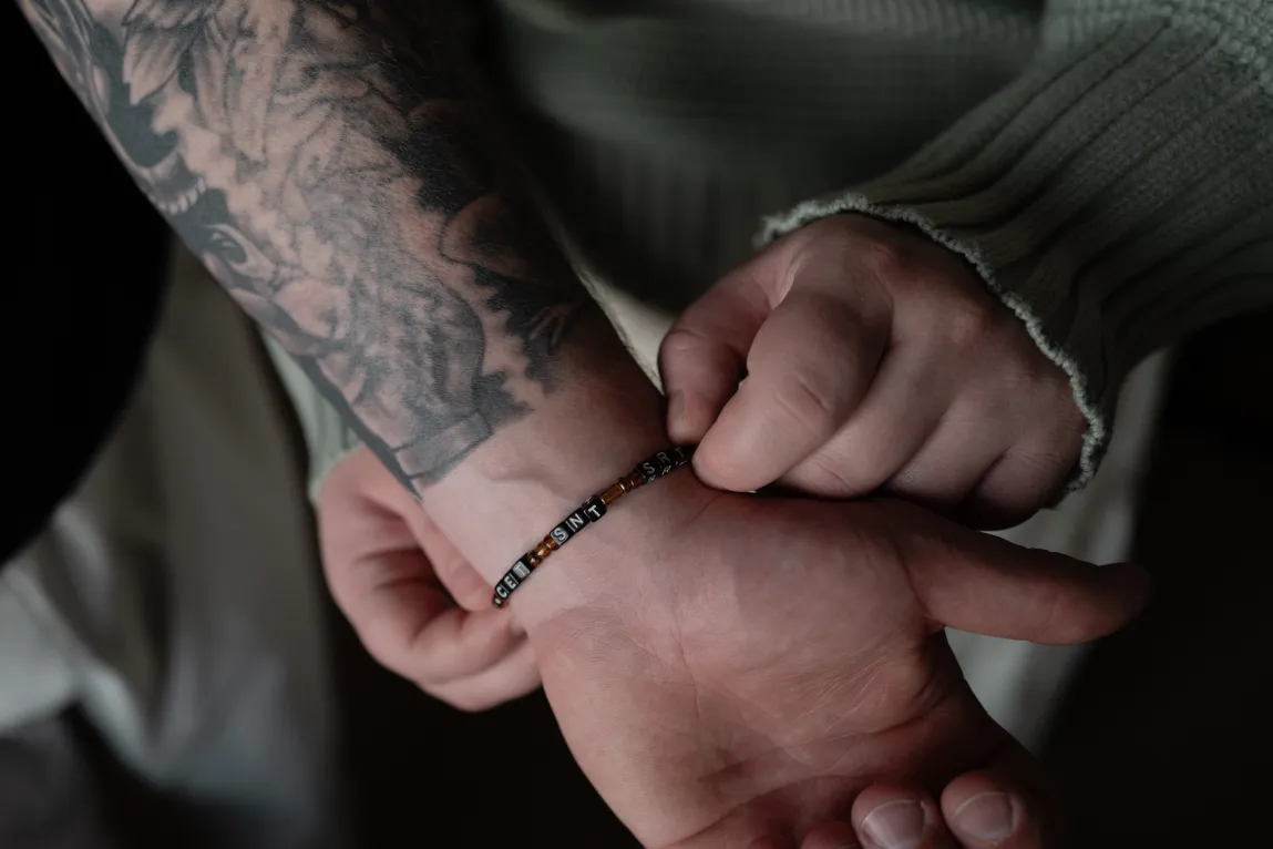 Gros plan de mains touchant un bracelet de perles porté au poignet, sous un bras tatoué.