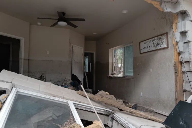 L’intérieur d’une maison montre de graves dégâts causés par les inondations, de hautes lignes de boue sur les murs, des parpaings effondrés et une pancarte encadrée indiquant « Donnez-le à Dieu et allez dormir ».