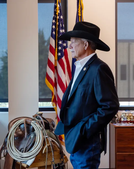 Un homme portant un chapeau de cowboy noir, un blazer bleu marine et un jean se tient de profil devant la caméra, devant un drapeau américain, une commode à médailles, une selle de cheval et une rangée de fenêtres.